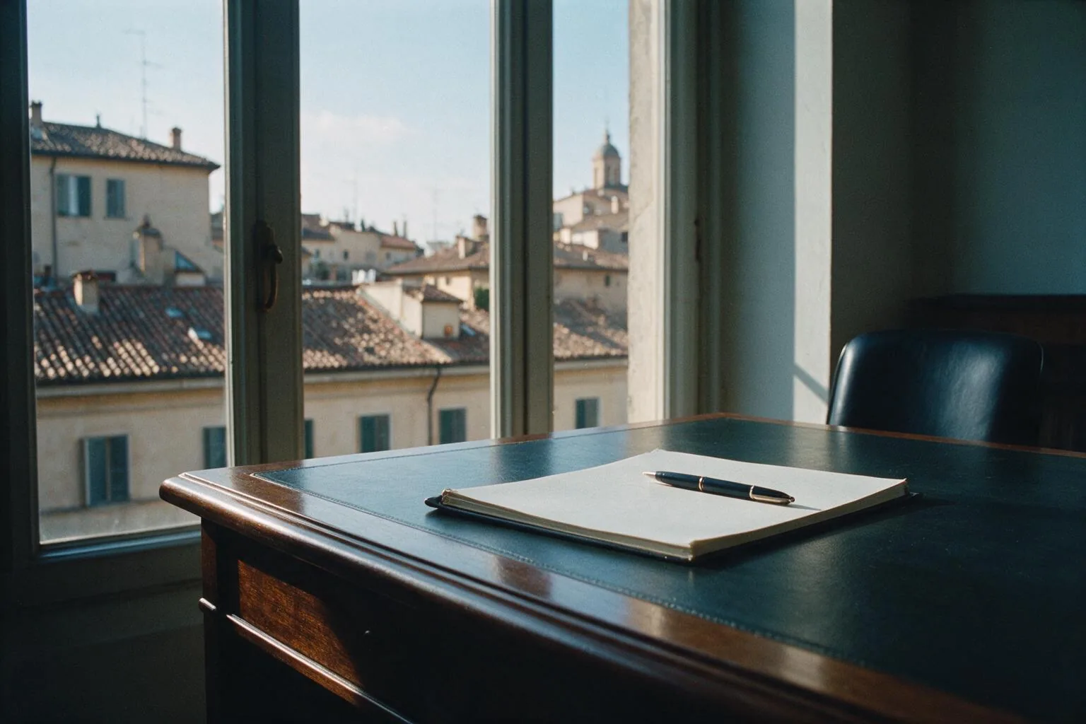 Una scrivania con un quaderno e una penna davanti a una grande finestra sui tetti di una città italiana