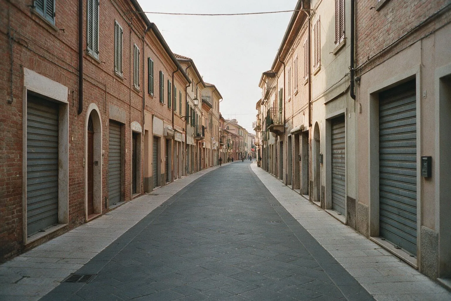Una strada di un centro storico emiliano, vuota, con case in mattoni e serrande abbassate nella luce di una mattina nuvolosa